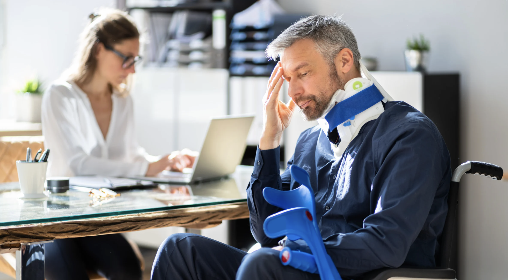 Man in neck brace and wheelchair, looking stressed in consultation with a woman working on a laptop, illustrating legal support for motorcycle accident victims in Texas.