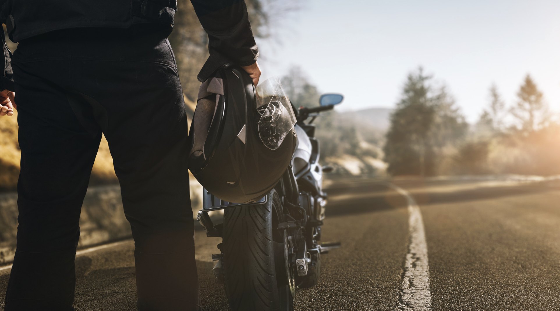 Motorcyclist holding a helmet while standing next to a motorcycle on a winding road, symbolizing motorcycle safety and legal considerations after accidents in Texas.