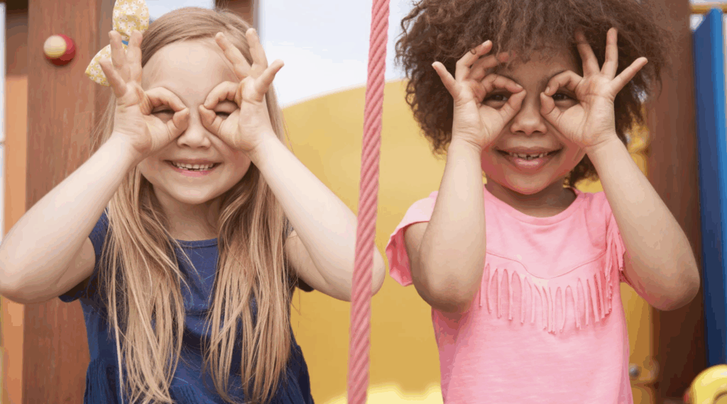 Two smiling girls at a playground making gestures with their hands to resemble glasses, highlighting joy and friendship in a playful setting, relevant to adoption themes of family and connection.