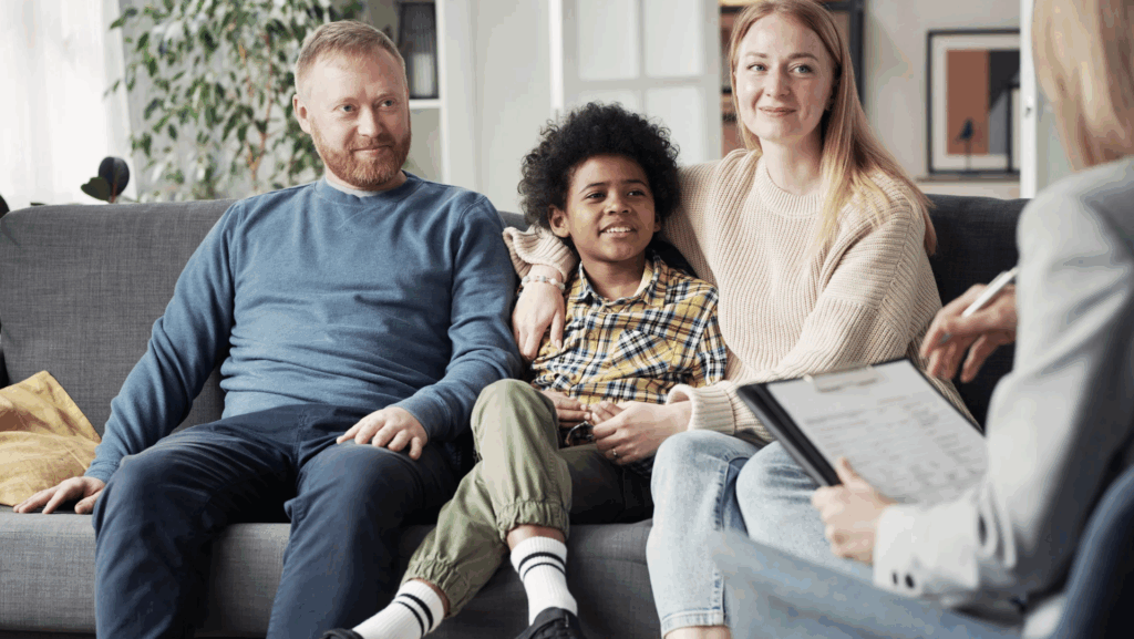 Screenshot 2025-09-30 at 2.29.13 PM Family sitting on a couch during an adoption consultation, smiling and engaged, with a child in a plaid shirt and an adult holding a clipboard.
