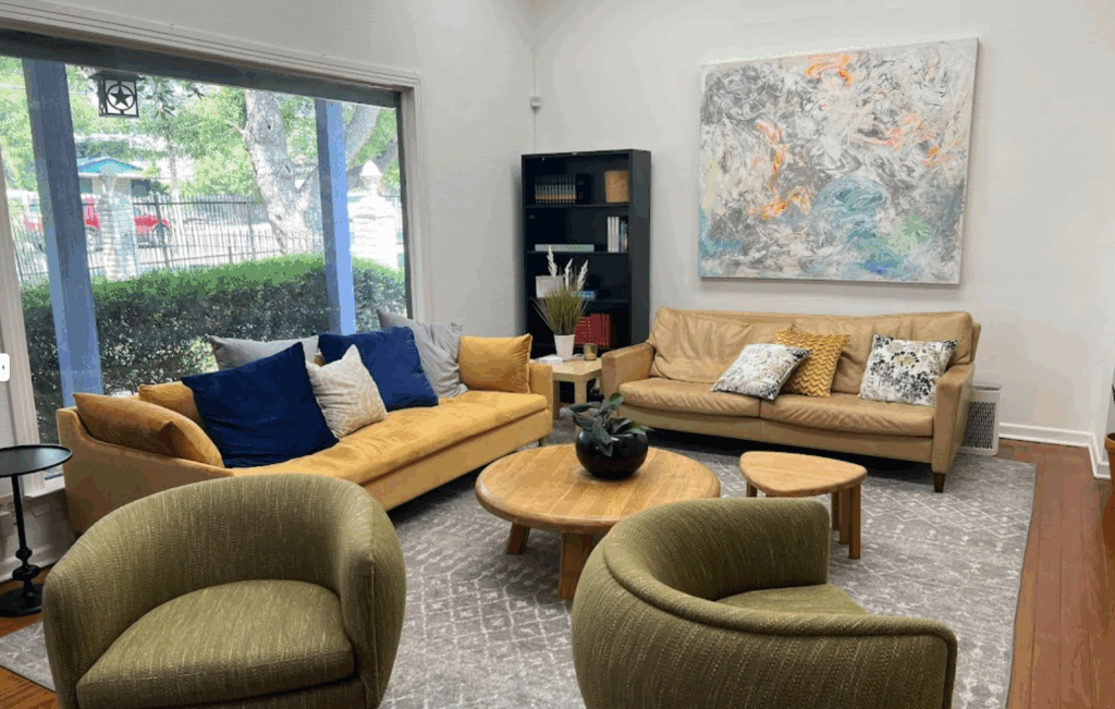 Cozy waiting area in the San Antonio office of the Law Office of Bryan Fagan, featuring yellow and brown sofas, decorative pillows, a round wooden coffee table, and a large abstract painting on the wall.