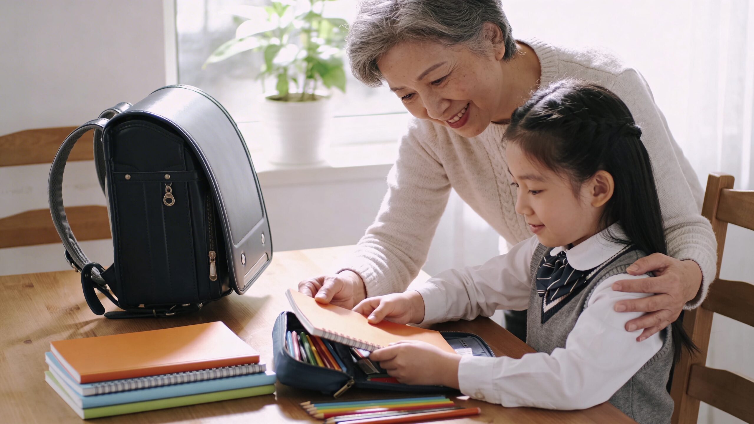 An elderly woman smiling while helping a young girl in a school uniform pack her pencil case.