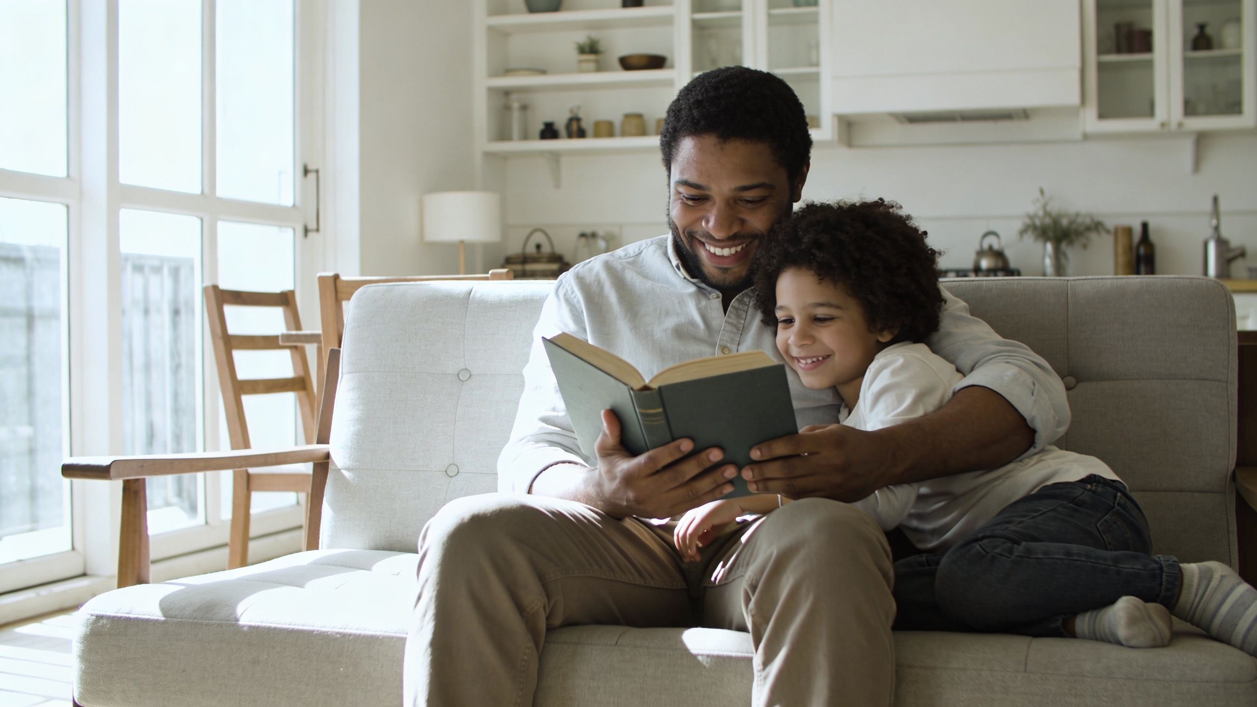 A father and son sitting on a sofa while happily reading a book together at home.